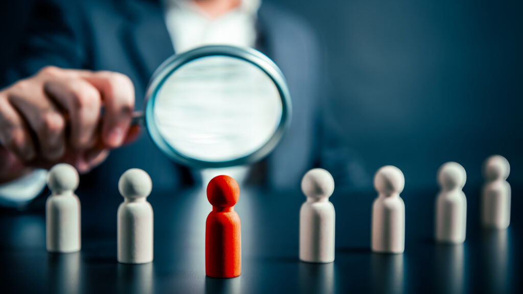 Person with magnifying glass verifying a red figure’s identiy among several white figures. 